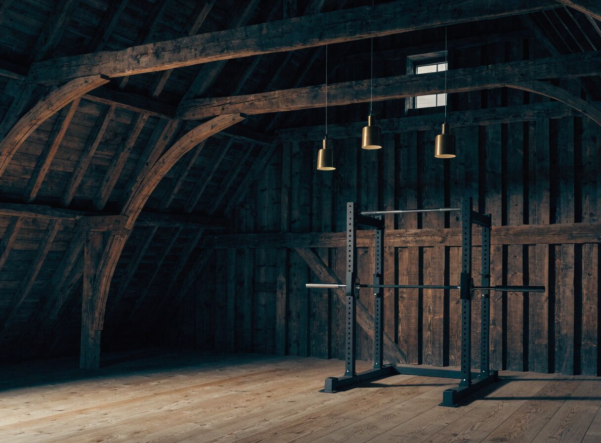 Concept render: a barn loft home gym with exposed timber framing, brass pendants, and a black half rack on an oak floor