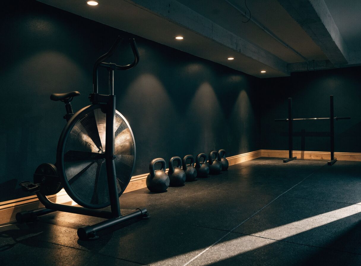 Concept render: a low-ceiling basement gym with an air bike, a row of kettlebells, and a short rack on rubber tile flooring
