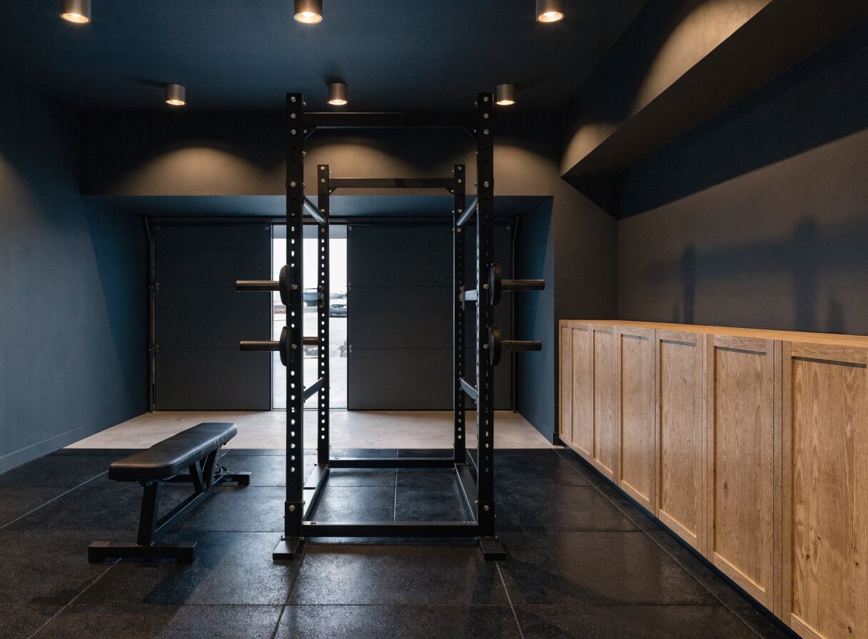 Concept render: a two-car garage converted to a family gym with a black power rack, flat bench, rubber tile floor, and oak cabinetry